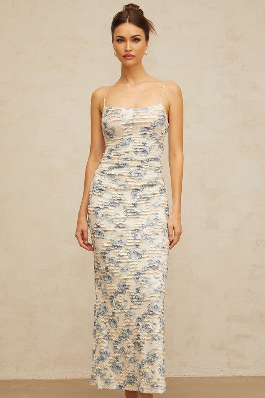 A woman wearing a long blue maxi dress with a textured floral print pattern standing against a plain background