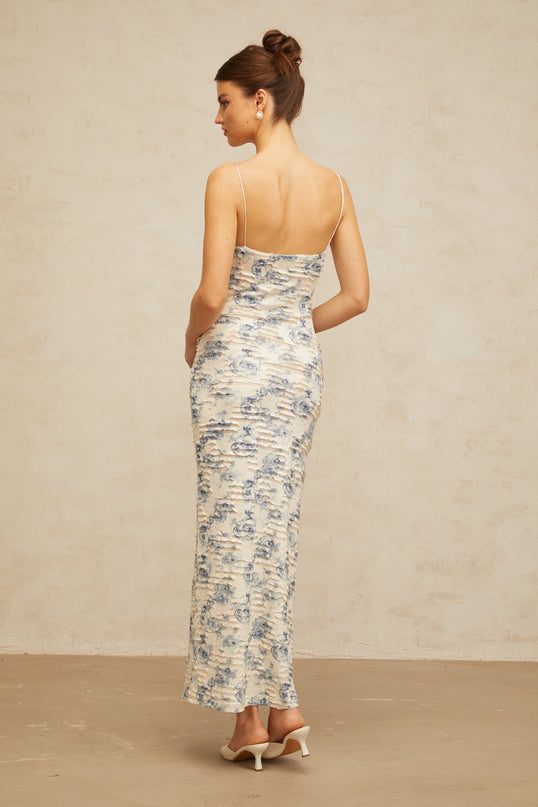 A woman wearing a long blue maxi dress with a textured floral print pattern The dress features a flowing silhouette and extends to the floor