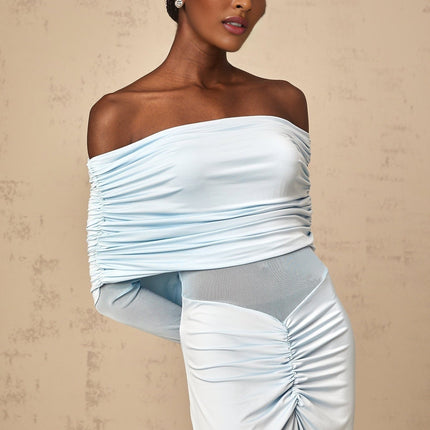 A woman wearing a long blue offshoulder dress with ruched detailing standing against a plain background