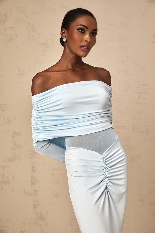 A woman wearing a long blue offshoulder dress with ruched detailing standing against a plain background