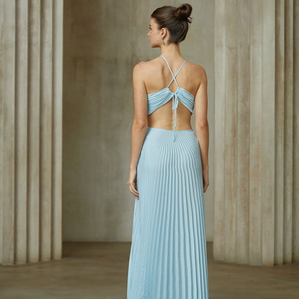 A woman wearing a long blue pleated maxi dress with a cutout detail on the waist standing against a plain background