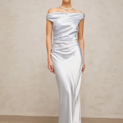 A woman wearing a long blue satin dress with offshoulder sleeves and ruched detailing standing against a plain background