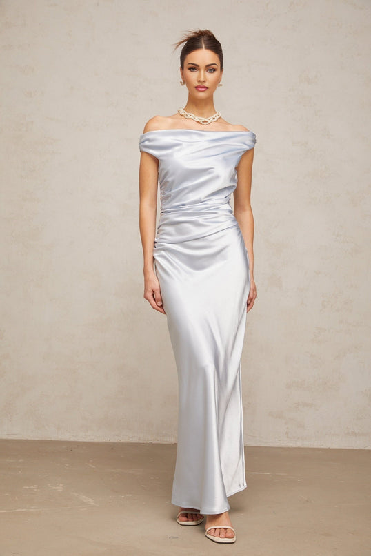 A woman wearing a long blue satin dress with offshoulder sleeves and ruched detailing standing against a plain background