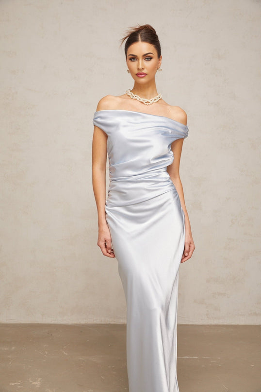 A woman wearing a long blue satin dress with offshoulder sleeves and ruched detailing standing in a relaxed pose