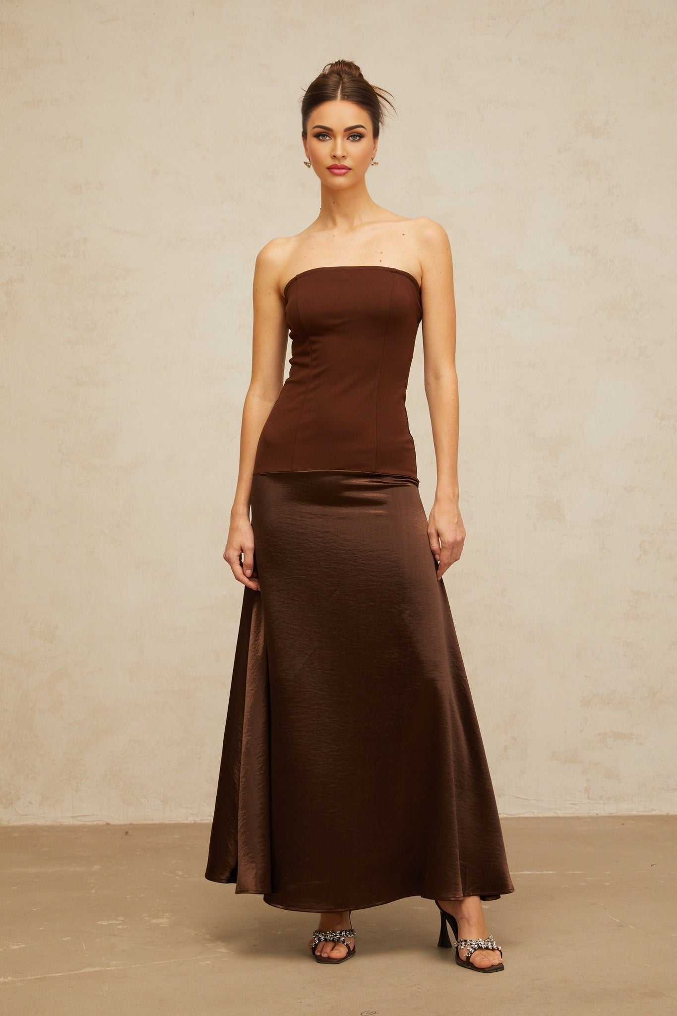 A woman wearing a long brown maxi dress with an offshoulder neckline and satin panel details