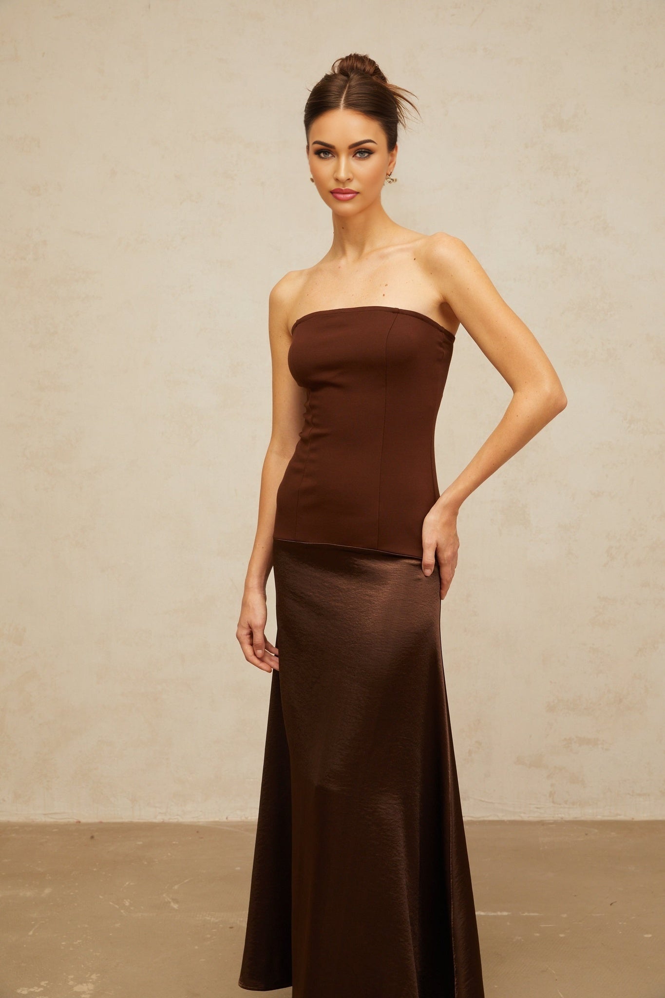 A woman wearing a long brown offshoulder dress with satin panels standing against a plain background