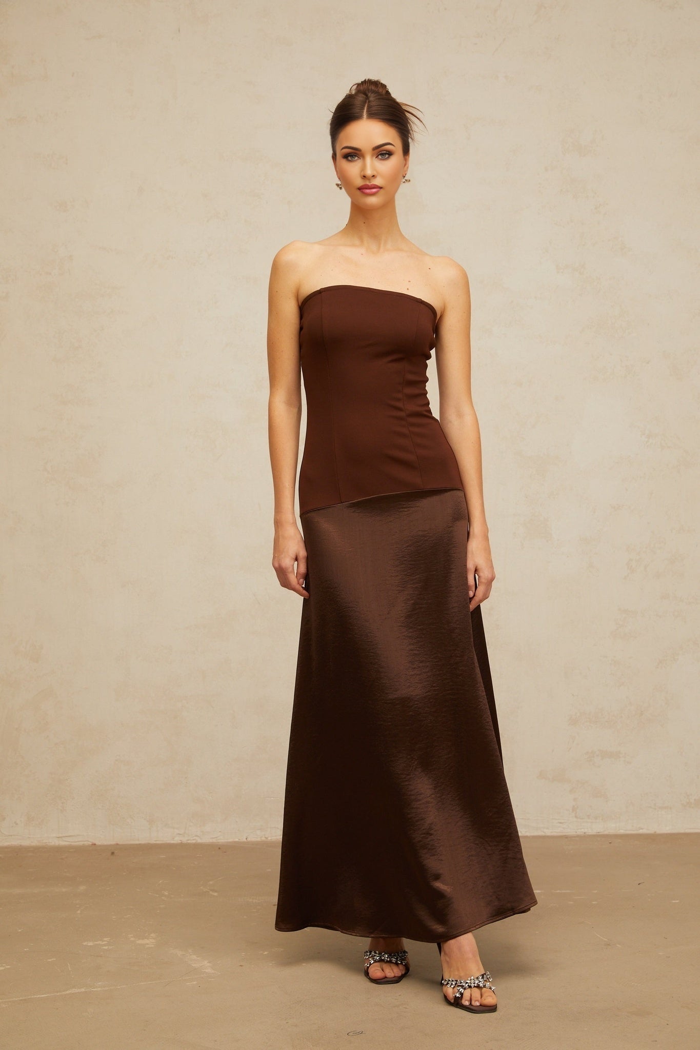 A woman wearing a long brown offshoulder dress with satin panels standing in a relaxed pose against a plain background