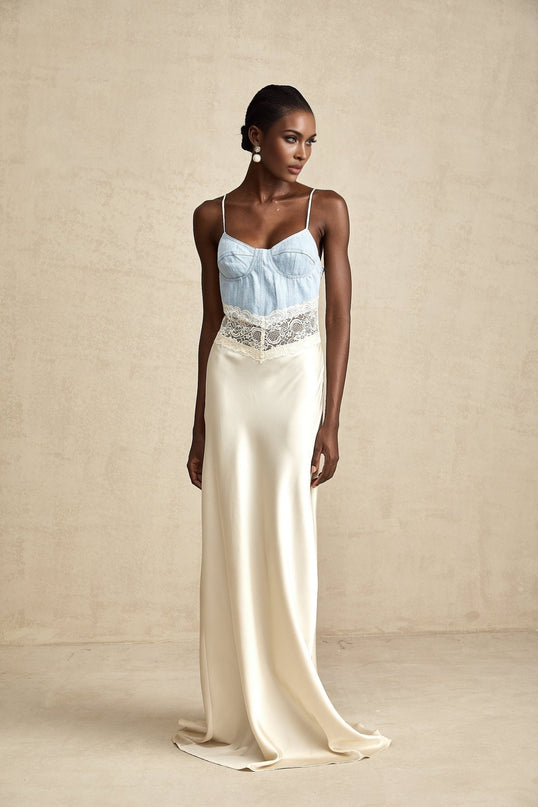 A woman wearing a long flowing Lisette maxi dress with lace trim and paneled design in a light color standing against a plain background