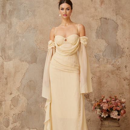 A woman wearing a long flowing neutral colored chiffon maxi dress with ruched detailing standing in a relaxed pose