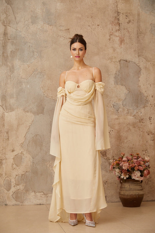 A woman wearing a long flowing neutral colored chiffon maxi dress with ruched detailing standing in a relaxed pose