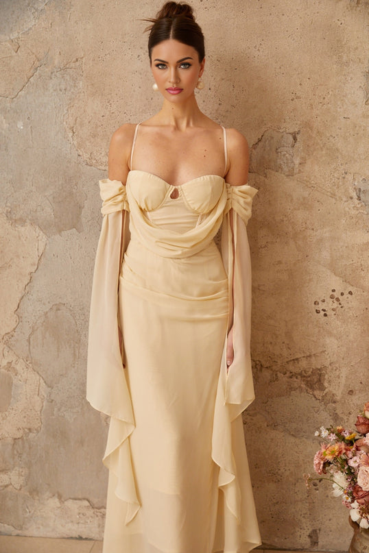 A woman wearing a long flowing neutral colored chiffon maxi dress with ruched detailing standing in a relaxed pose