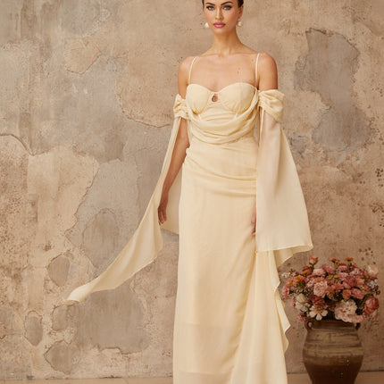 A woman wearing a long flowing neutralcolored chiffon maxi dress with ruched detailing standing in a relaxed pose