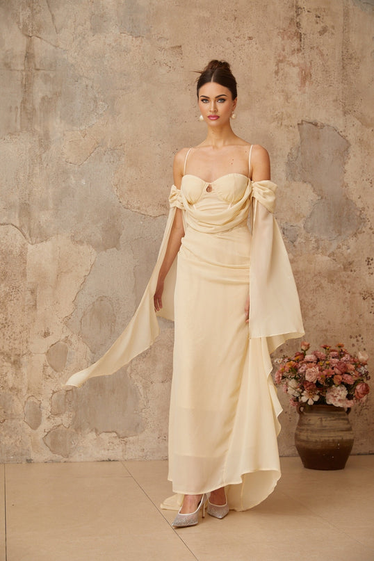 A woman wearing a long flowing neutralcolored chiffon maxi dress with ruched detailing standing in a relaxed pose