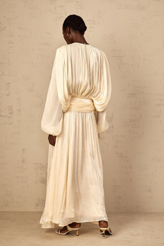 A woman wearing a long flowing pleated maxi dress with ruffled details in a neutral beige color standing in a relaxed pose