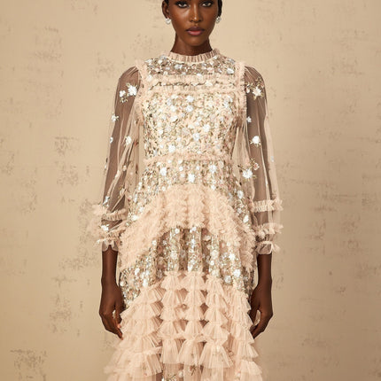 A woman wearing a long flowy maxi dress with floral embroidery and tiered tulle skirt in a soft pastel color standing against a plain background