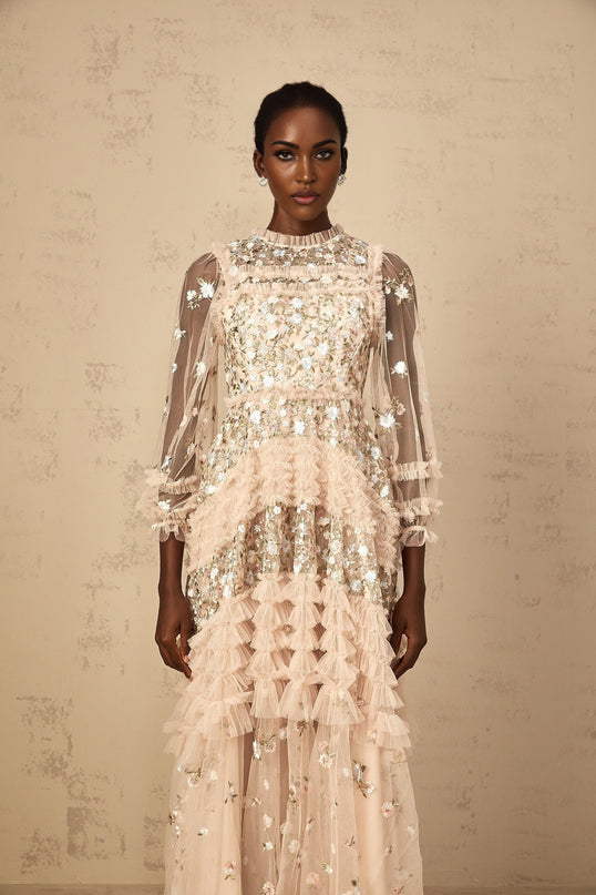 A woman wearing a long flowy maxi dress with floral embroidery and tiered tulle skirt in a soft pastel color standing against a plain background