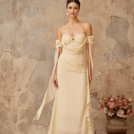 A woman wearing a long flowy neutral colored chiffon maxi dress with ruched detailing standing in a relaxed pose