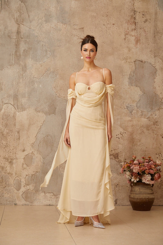 A woman wearing a long flowy neutral colored chiffon maxi dress with ruched detailing standing in a relaxed pose