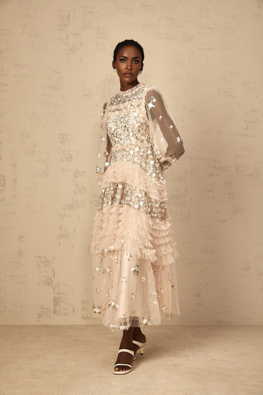 A woman wearing a long flowy tulle maxi dress with delicate floral embroidery and tiered layers in soft pastel colors standing in an elegant pose