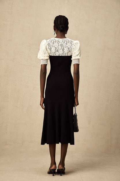 A woman wearing a long knit midi dress with lace trim details along the neckline and hem standing in a relaxed pose against a plain background