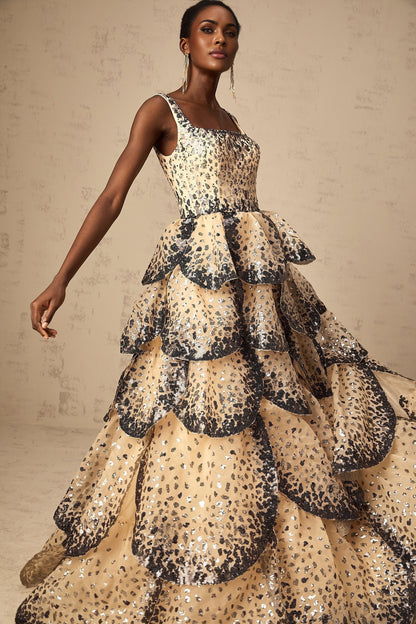 A woman wearing a long maxi dress with tiered layers and shimmering sequin details in a petal design standing in an elegant pose
