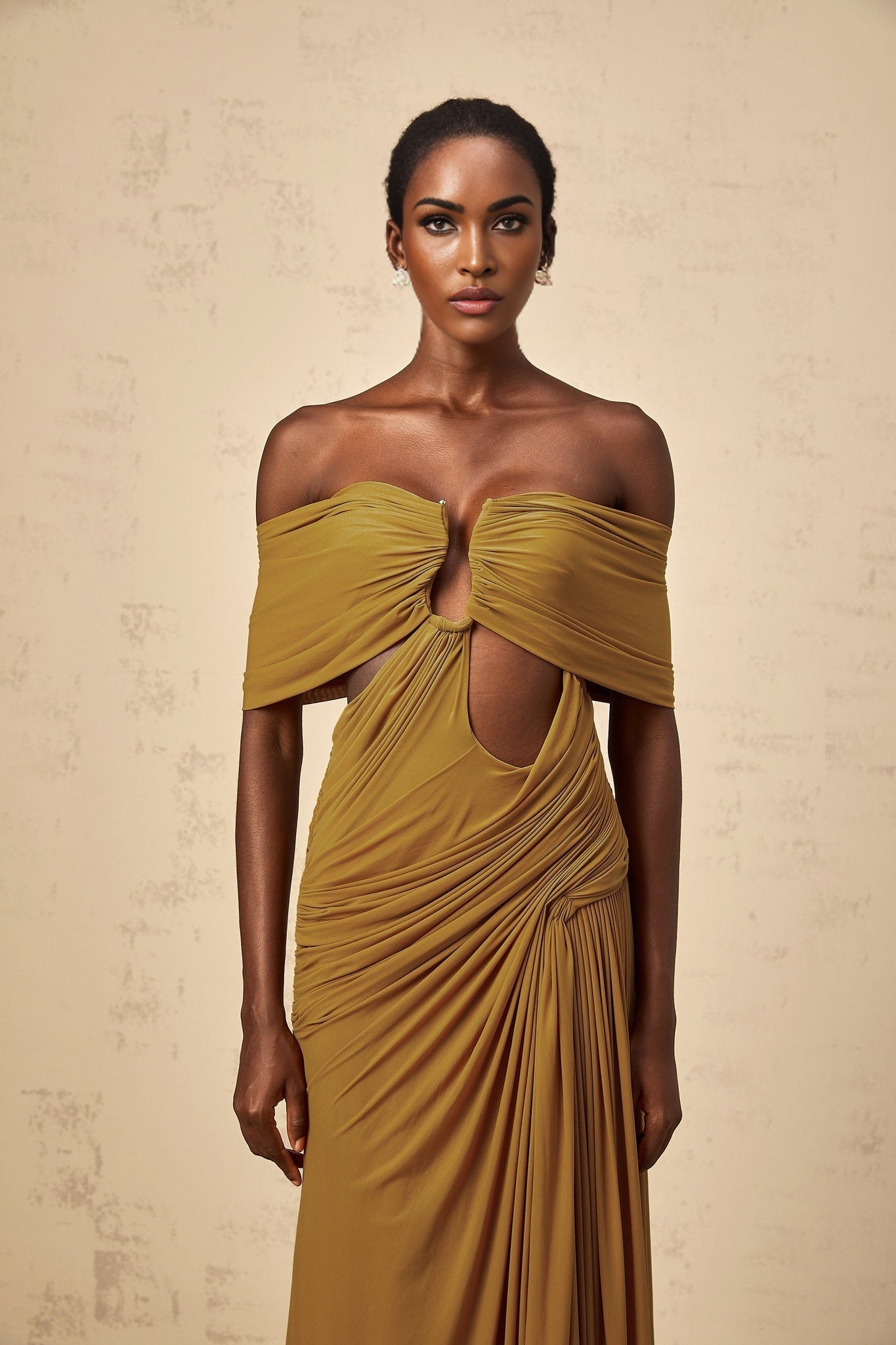 A woman wearing a long ocher colored dress with offshoulder design and ruched details The dress features cutouts and falls to a maxi length