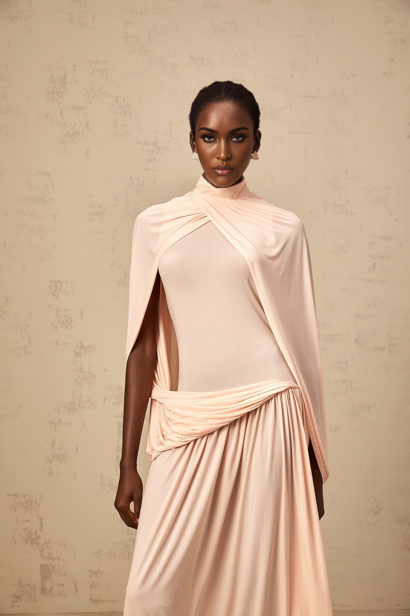 A woman wearing a long pink maxi dress with a flowing cape design and asymmetrical hemline
