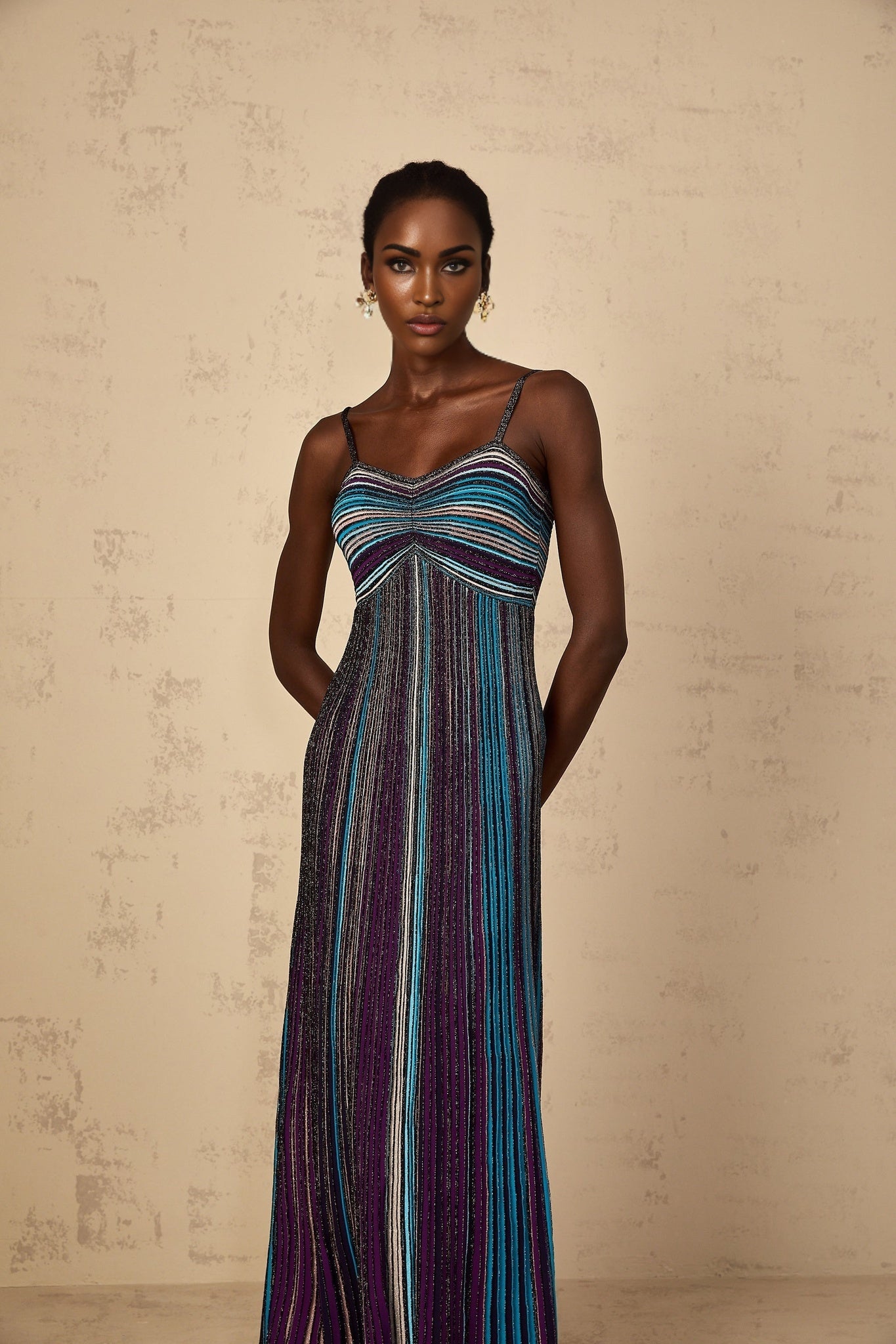 A woman wearing a long purple maxi dress with horizontal sequin stripes knitted into the fabric the dress has a form fitting silhouette and extends to the floor