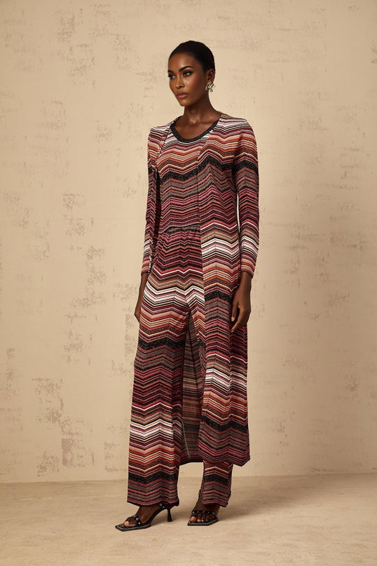 A woman wearing a long red cardigan with white stripes open over a white top and blue jeans standing against a plain background