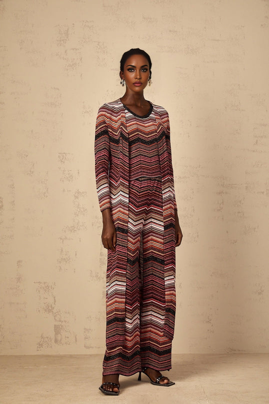 A woman wearing a long red cardigan with white stripes standing against a plain background The cardigan has a relaxed fit and reaches below the knees