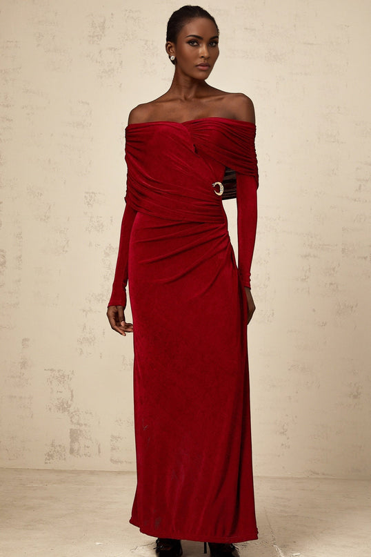 A woman wearing a long red velvet dress with ruched detailing and a fitted silhouette standing against a plain background