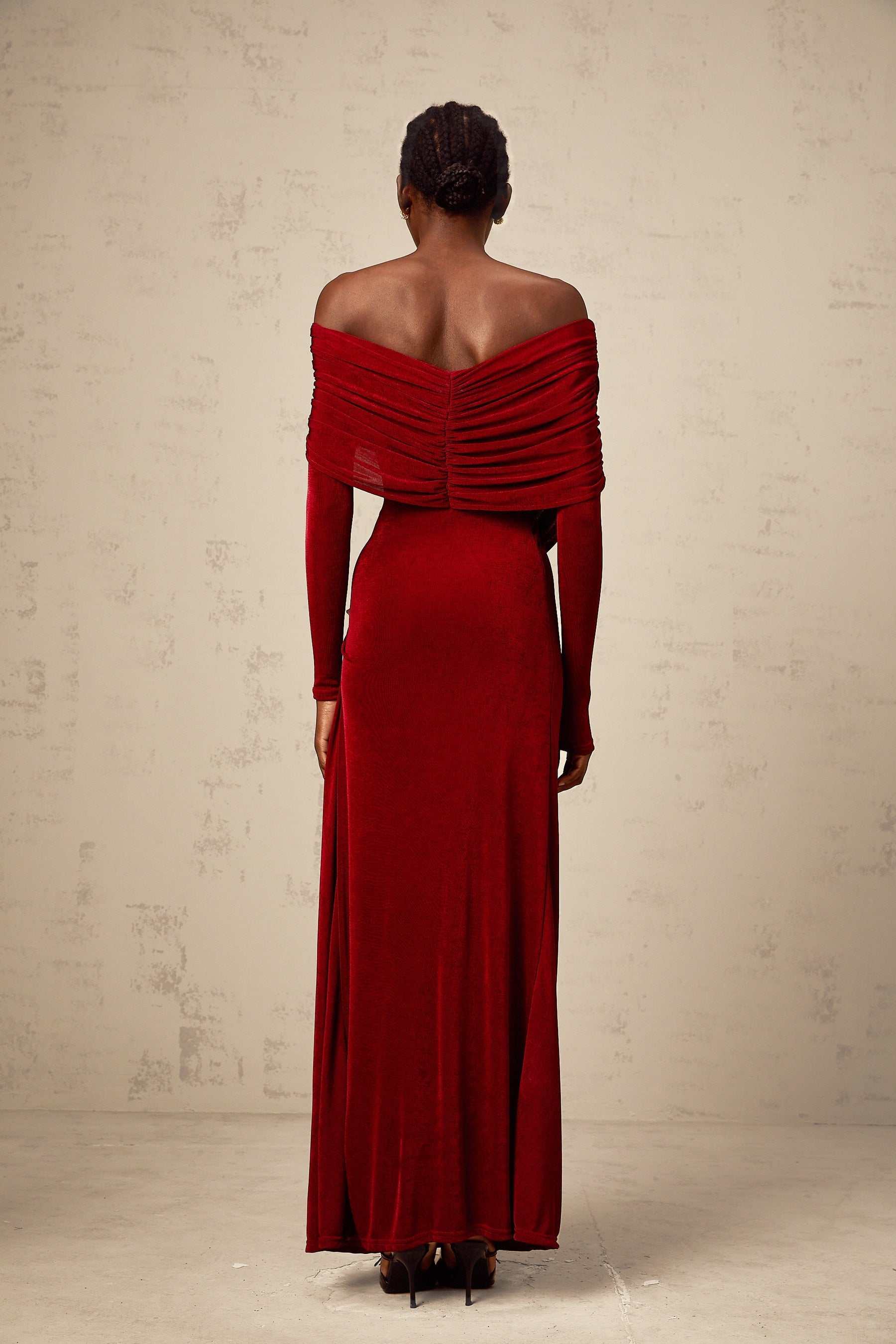 A woman wearing a long red velvet dress with ruched detailing and a fitted silhouette standing against a plain background