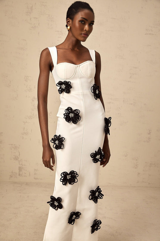 A woman wearing a long white formfitting dress with floral applique details standing against a plain background