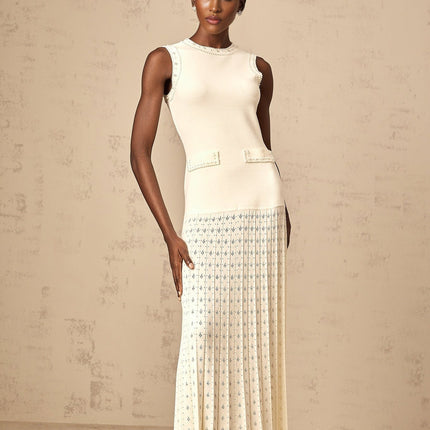 A woman wearing a long white knitted maxi dress with faux pearl embellishments standing in a neutral background