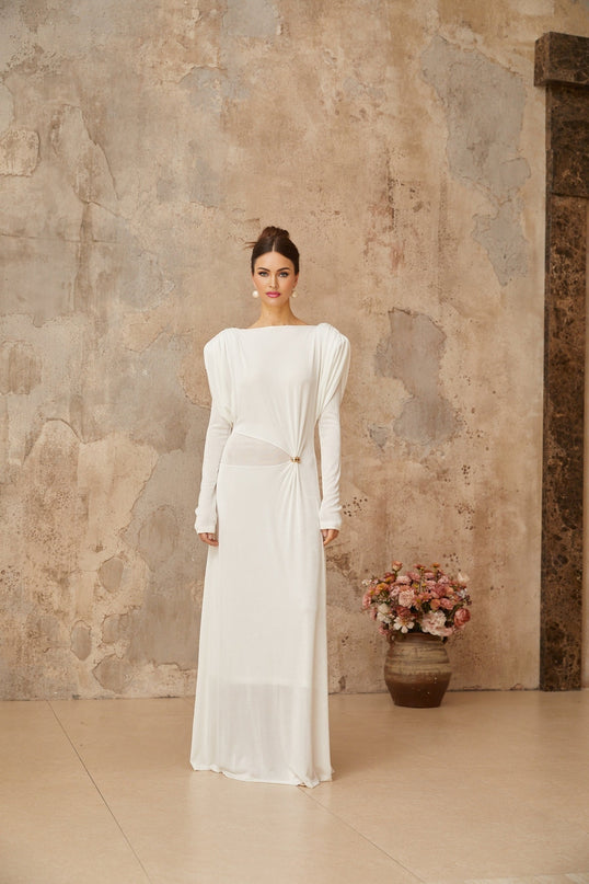 A woman wearing a long white maxi dress with draped and shirred details standing against a plain background