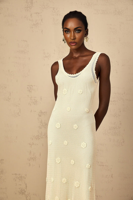 A woman wearing a long white maxi dress with floral embroidery and crochet knit details standing in a garden setting