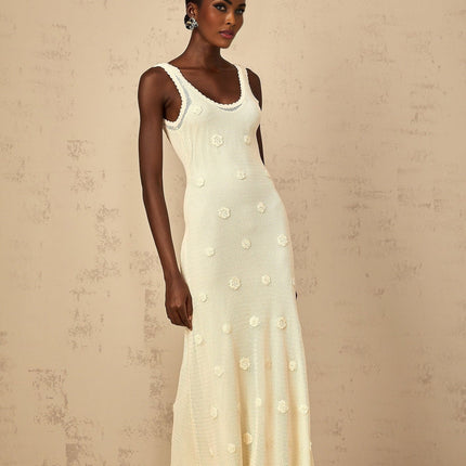 A woman wearing a long white maxi dress with floral embroidery and crochet knit details standing in a garden setting