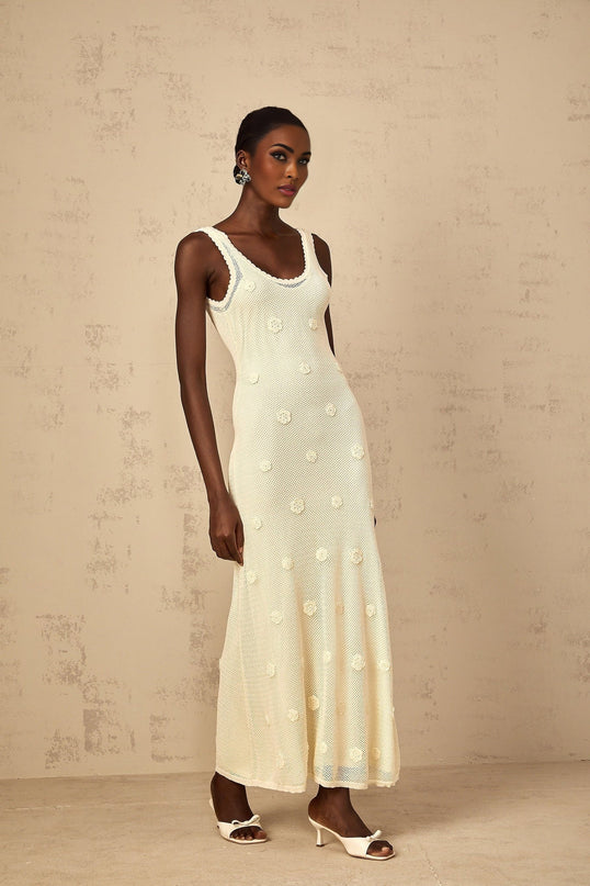 A woman wearing a long white maxi dress with floral embroidery and crochet knit details standing in a garden setting