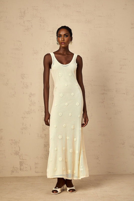 A woman wearing a long white maxi dress with floral embroidery and crochet knit details standing in an outdoor setting