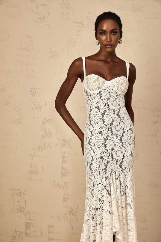 A woman wearing a long white maxi dress with floral lace details and a bustier style top The dress has a fitted bodice and flows into a full length skirt