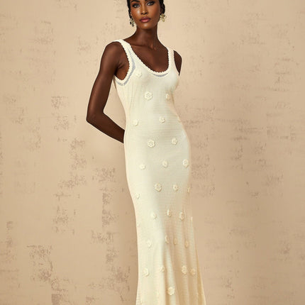 A woman wearing a long white maxi dress with intricate floral embroidery and crochet knit details standing in a garden setting