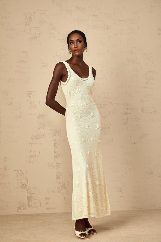 A woman wearing a long white maxi dress with intricate floral embroidery and crochet knit details standing in a garden setting