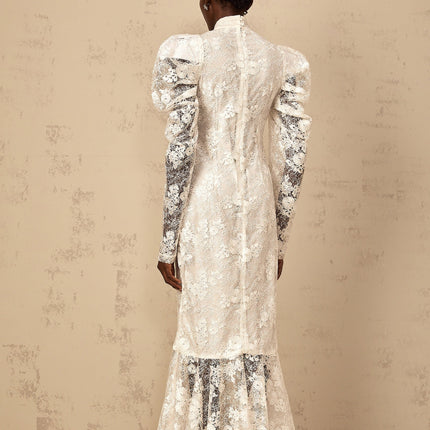 A woman wearing a long white mermaid style dress with floral lace pattern and fitted bodice standing against a plain background