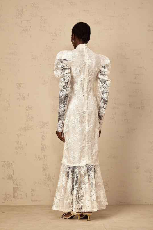 A woman wearing a long white mermaid style dress with floral lace pattern and fitted bodice standing against a plain background