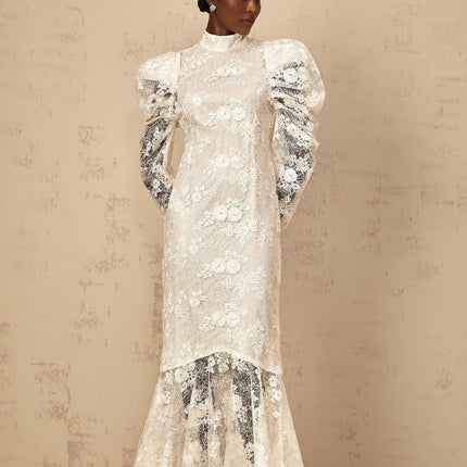 A woman wearing a long white mermaid style dress with floral lace pattern standing in an elegant pose