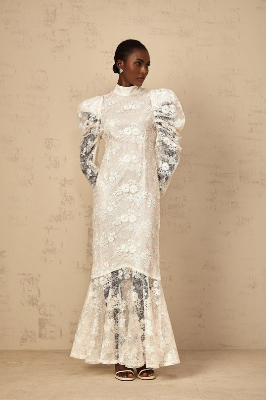A woman wearing a long white mermaid style dress with floral lace pattern standing in an elegant pose