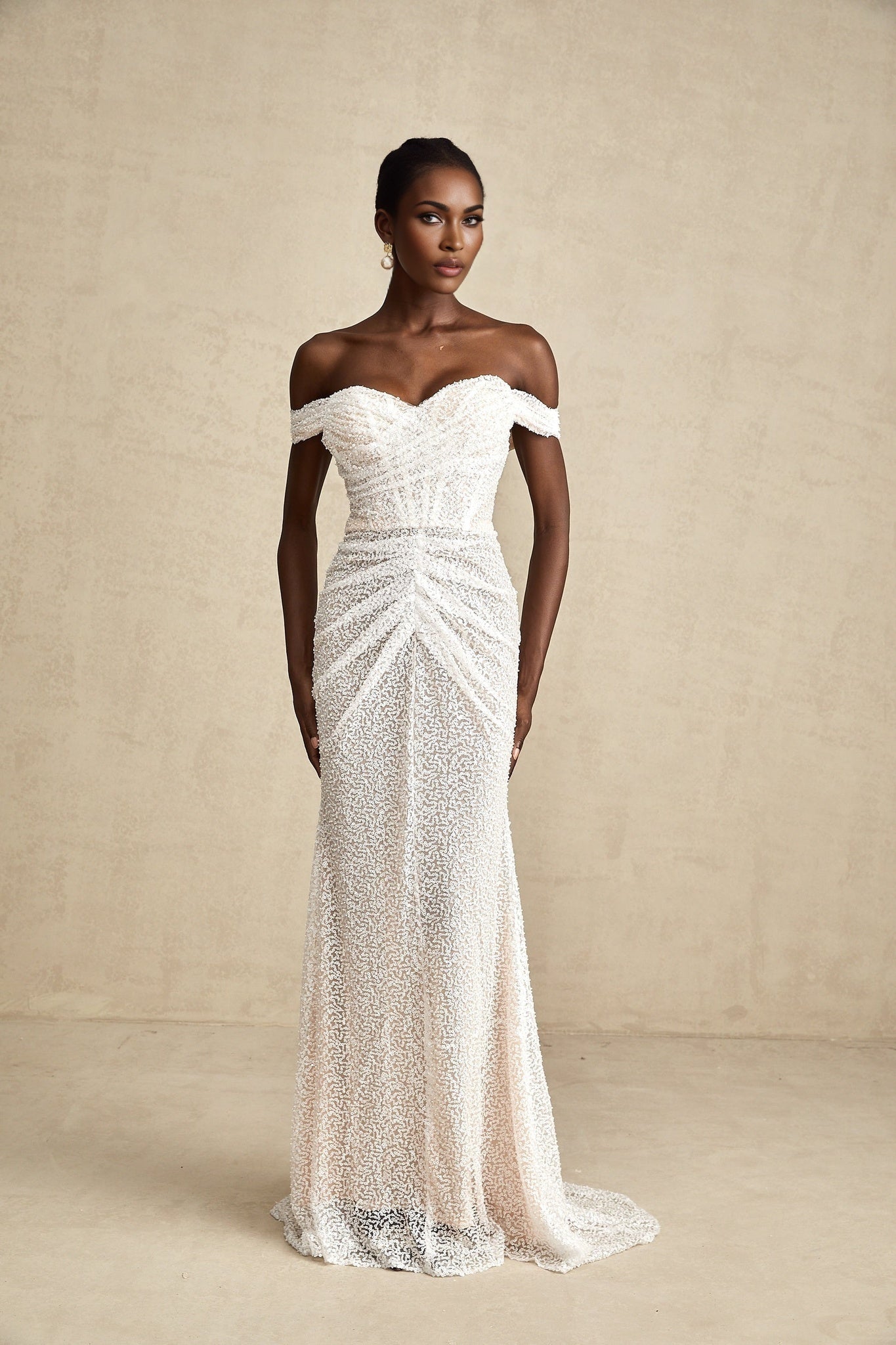 A woman wearing a long white offshoulder maxi dress with embellishments standing in a relaxed pose against a plain background