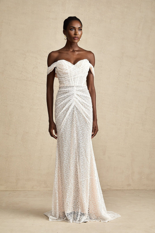 A woman wearing a long white offshoulder maxi dress with embellishments standing in a relaxed pose