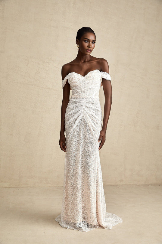 A woman wearing a long white offshoulder maxi dress with embellishments standing in a relaxed pose