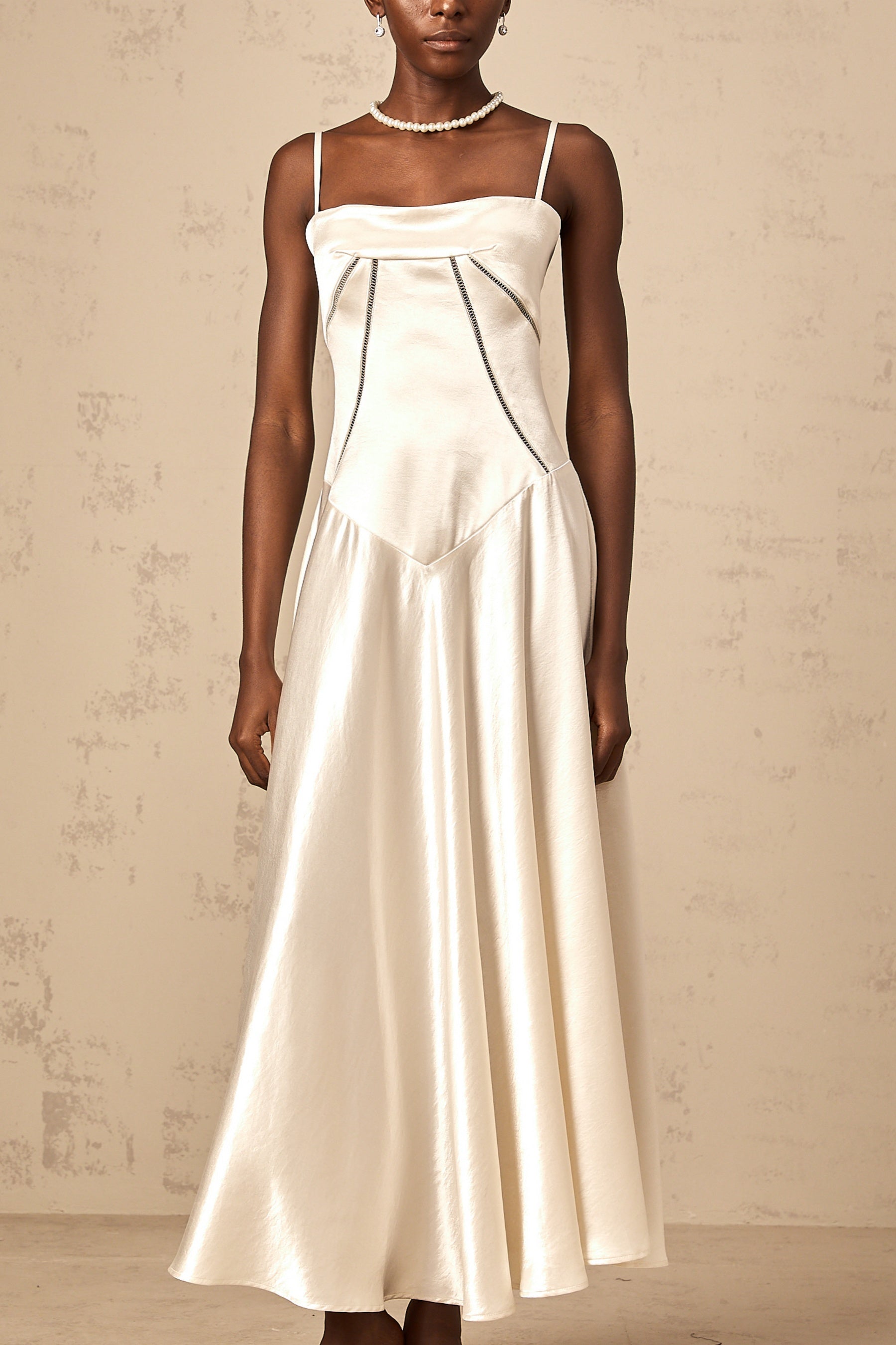 A woman wearing a long white satin dress with a flared skirt and fitted bodice standing against a plain background